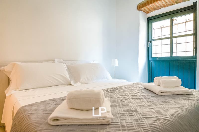 Spacious bedroom with white walls, large windows, wooden ceiling beams, and a blue wooden door. The room features a comfortable bed with white bedding, a patterned blanket, and plush white towels. The overall atmosphere is bright and airy.