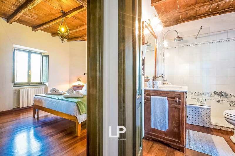 The image shows a bedroom with a wooden ceiling and a chandelier, and an adjoining bathroom with a vanity and tiled walls. The bedroom has a bed with a blue and green bedspread, and the bathroom has a wooden vanity and towels.
