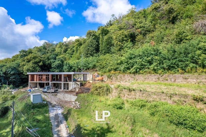 Under construction house on the side of a hill, with a sloped terrain in the foreground, surrounded by trees and greenery, with machinery and vehicles on site.