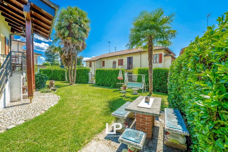 Two houses, garden with table and bench, palm trees, well-maintained lawn, hedges, metal fence, patio, covered porch, staircase, clear sky