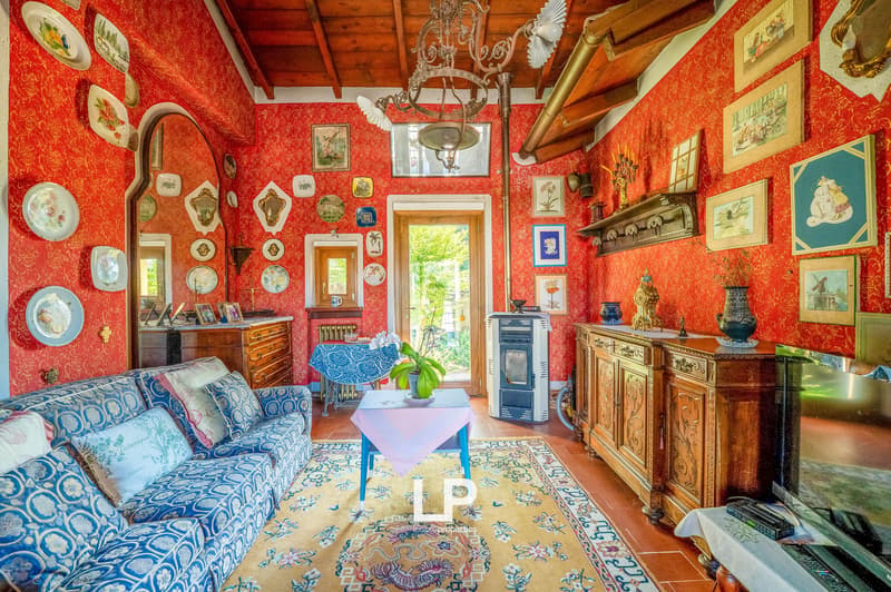 red wallpaper, ornate ceiling chandelier, sofa, rug, table with flower, side table, television, mirror, side table with candles, fireplace, picture frames, cabinet, radiator, large mirror, window with curtains