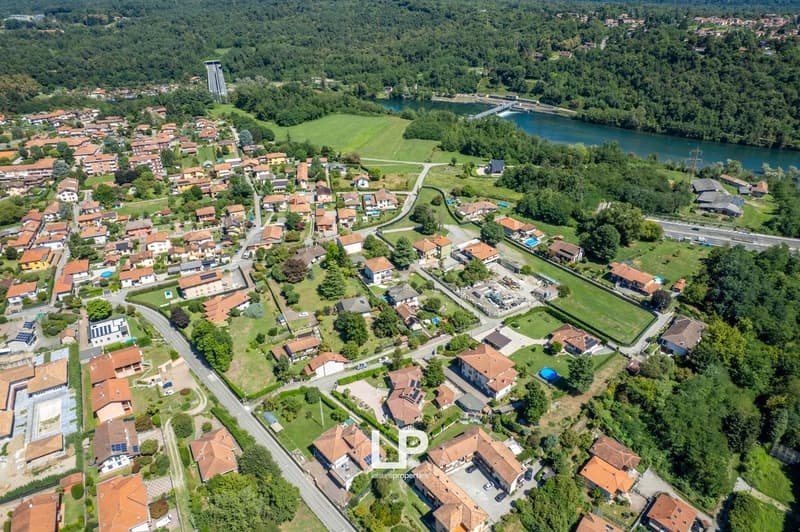 Suburban neighborhood, multi-story houses, red roofs, greenery, some houses have solar panels, swimming pools, river visible in the distance, trees, well-kept areas