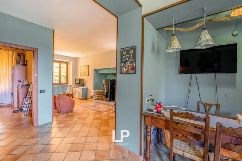 Dining room, large tiled floor, wooden table and chairs, hanging lamps, TV, fireplace, framed poster, shelves, cabinet, ladder, small armchair, curtains, radiator