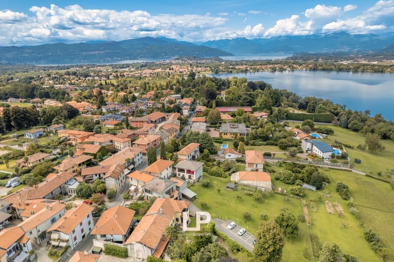 Small town with houses clustered, surrounded by green trees and plants, body of water in the background.