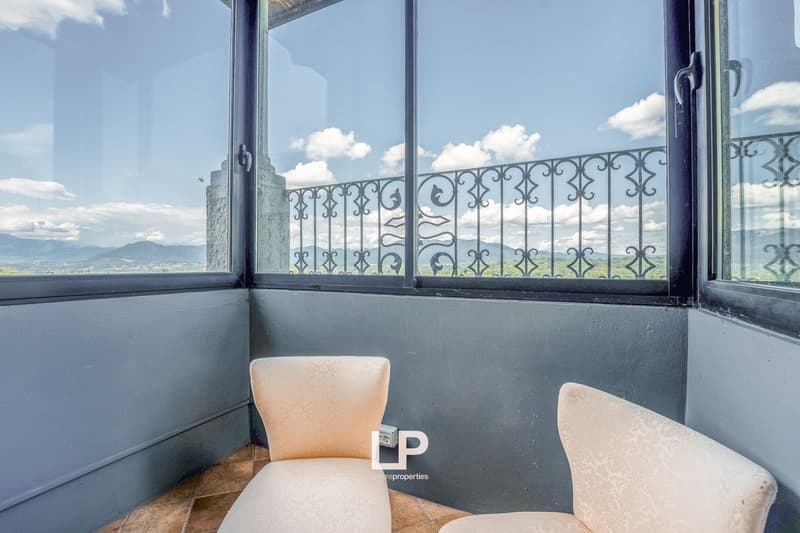 Gray walls, tiled floor, two beige upholstered chairs, black metal railing with intricate design, large window providing a view of mountains and sky.