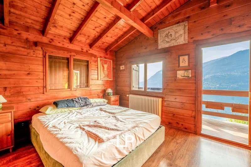 Bedroom with wooden walls, wooden ceiling beams, bed with white sheets, nightstand with lamp, radiator, sliding glass doors leading to a balcony with a lake view, picture frames on the wall