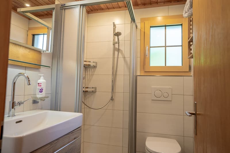 modern bathroom with white tiles, glass door shower, white sink, silver faucet, mirror, small shelf, toilet, towel rack, power outlet