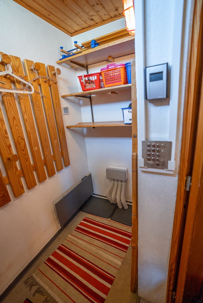 Storage room with wooden panels for hanging clothes, shelves with storage baskets, a white mat on the floor, an intercom system, and a white wall.