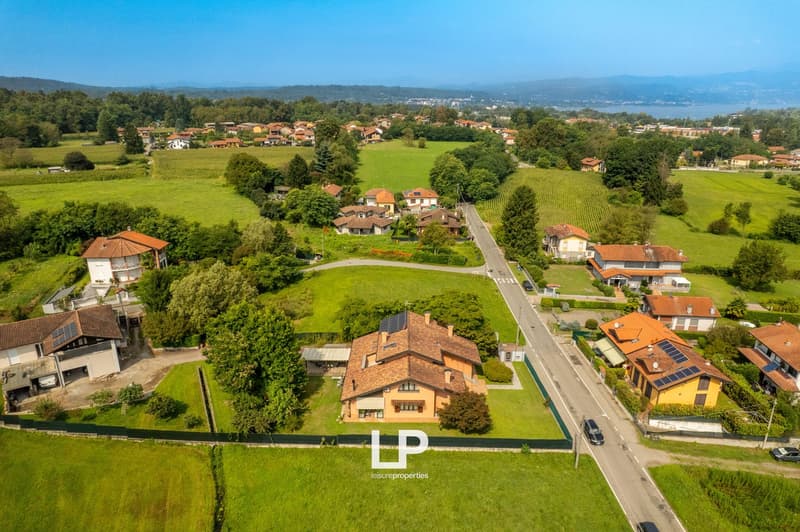 Countryside house with solar panels, surrounded by green fields, roads, and other houses. Mountain and lake in the distance.