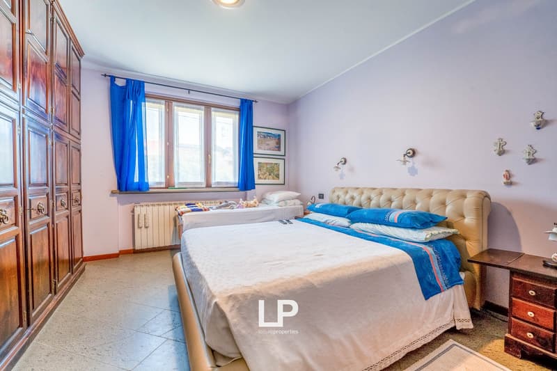 Large bed, second smaller bed, windows with blue curtains, two picture frames, wall art, wooden wardrobe, nightstand, radiator, tile floor.