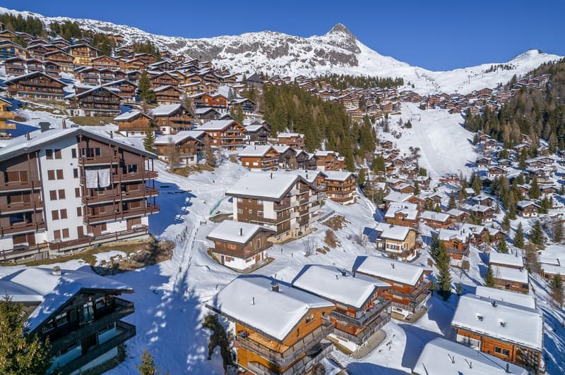 Aerial view of numerous chalet-style houses on a mountain, all surrounded by snow, with a ski slope and trees in the background.