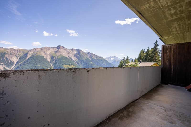 concrete wall, concrete floor, open wooden door, expansive view of mountain range, clear blue sky with clouds, distant house under trees