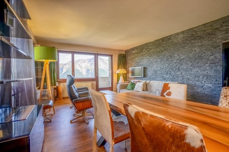 Open floor plan, wooden table and chairs, leather chairs, sofa with pillows, TV on the wall, glass window with a mountain view, floor lamp, wooden shelves with a glass top, and a desk with a lamp.