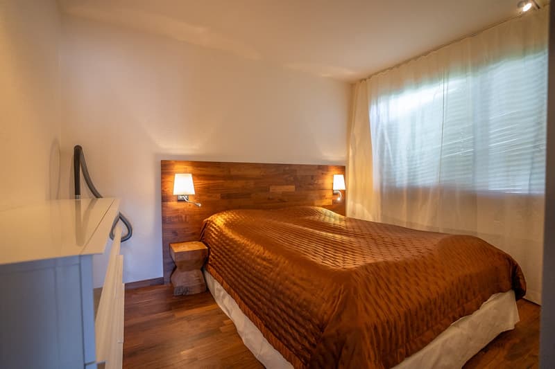 wooden headboard, white walls, hardwood floor, window with white curtains, bedside tables with lamps, large bed with orange comforter