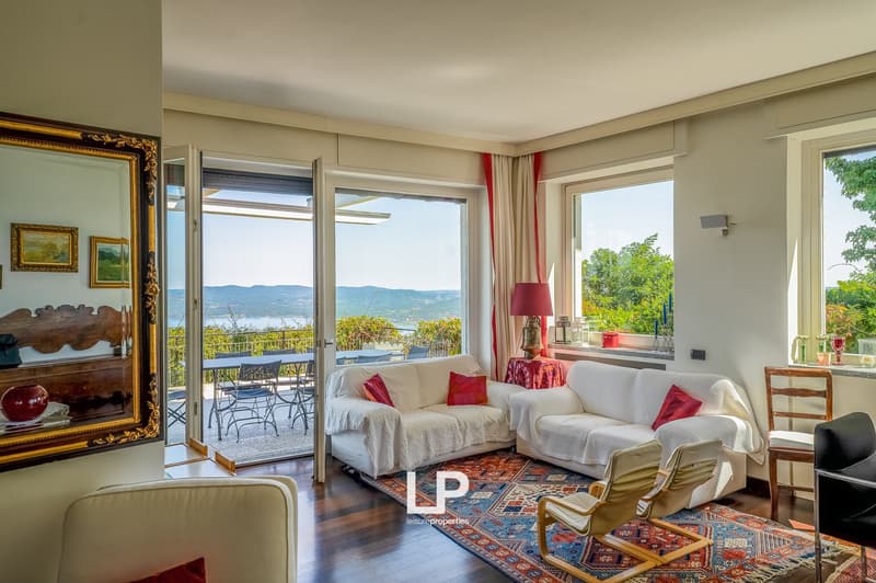 Spacious living room with double sofa, coffee table, carpet, two white couches with red cushions, mirror, and large windows with balcony. Table and chairs on the balcony.