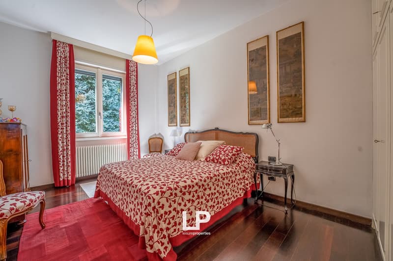 king size bed, red bedding, two nightstands, wooden cabinet, red curtains, wooden floor, radiator, window, lamps