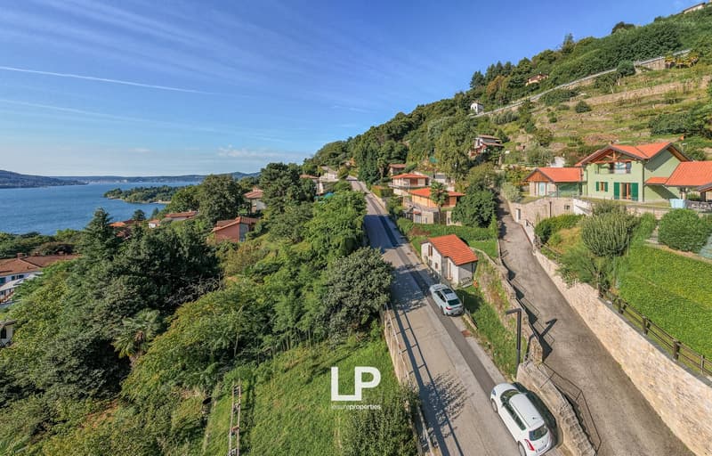 Terraced houses with red roofs, one car in driveway, road with cars, view of a bay with a small island, green hillside with plants and trees