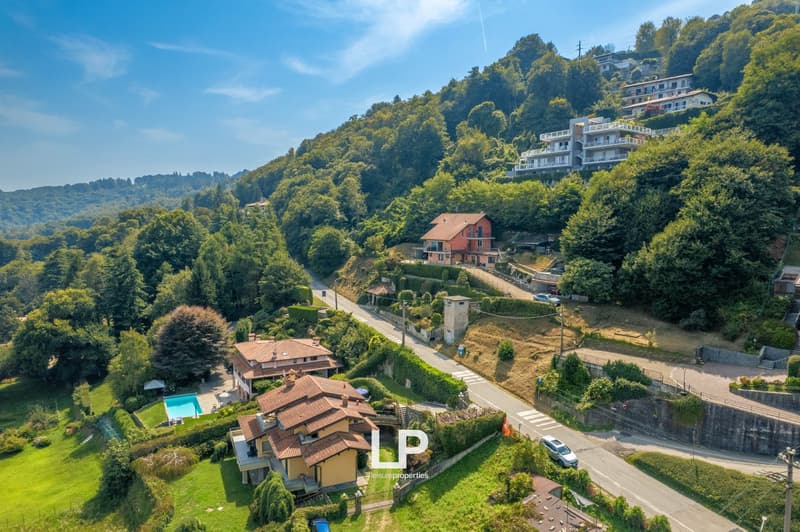 Two houses with swimming pool, nestled on a hillside with lush greenery, mountain backdrop, driveway leading to houses, blue car on road, clear blue sky, residential area with distant mountains