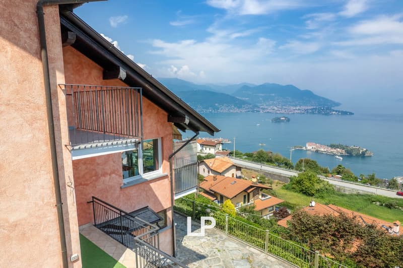 Facade of the house with a balcony, large windows, surrounded by green hills and a stunning view of the lake