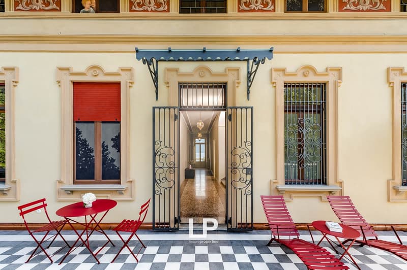 A beige building with an arched entrance and a black and white checkerboard floor in the hallway. It has multiple large windows with red shutters and a few decorative red chairs.