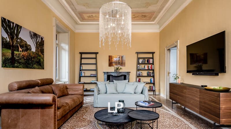 A living room with two couches, shelves with books, a fireplace, a table with magazines, TV mounted on the wall, and a chandelier