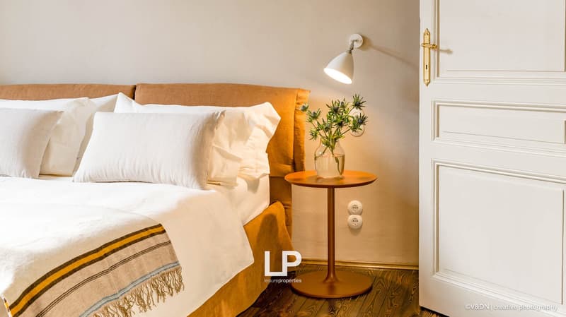 A bed with pillows and a blanket, a table with flowers, a wall lamp, and a white door