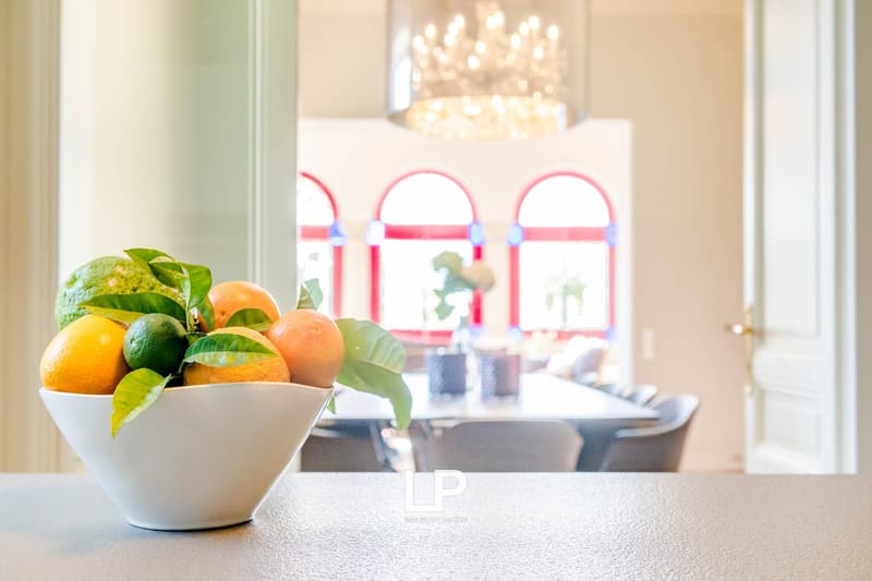 Bowl of fruits on a table in front of arched windows and a dining table with chairs.