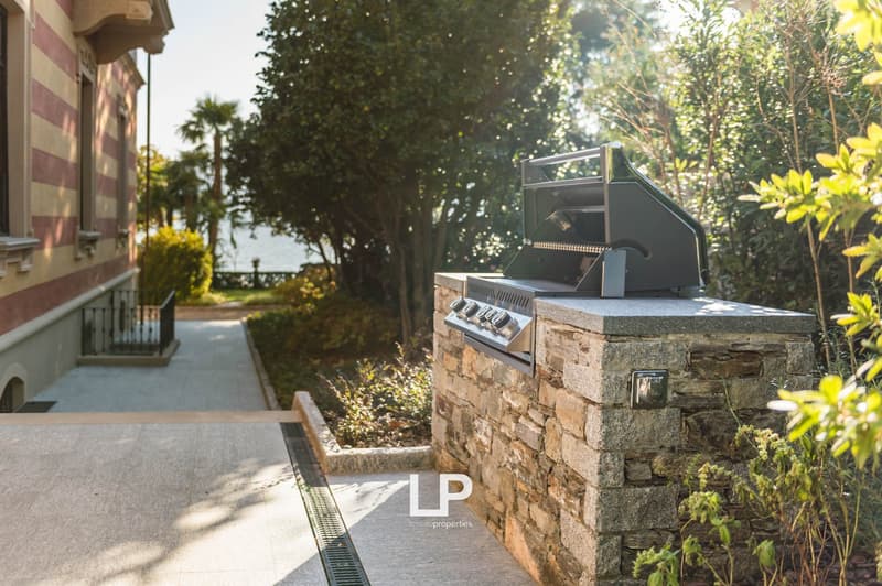 Outdoor area with a grill and stone wall, pathway leading to the grill, plants and trees, building visible on the left