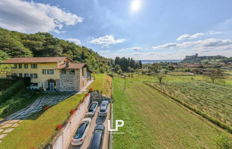 Stone house with red tiled roof, 4 cars parked in front, located on a hill with a vineyard and fields in the background.