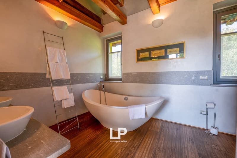 Large white bathtub, white sinks, white towels on towel rack, picture frame, windows, wooden floors, wall mounted lights