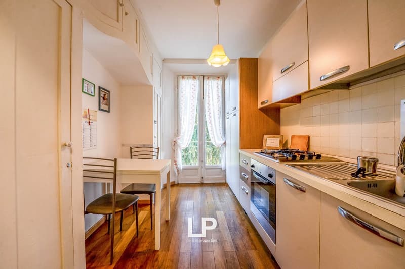 Open kitchen with white countertop, wooden floor, gas stove, sink, wooden cabinets, table and chairs.