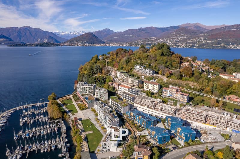 Aerial view of a hillside community with waterfront properties, featuring modern apartment buildings, a marina with docked boats, expansive lake views, and a mountainous backdrop.