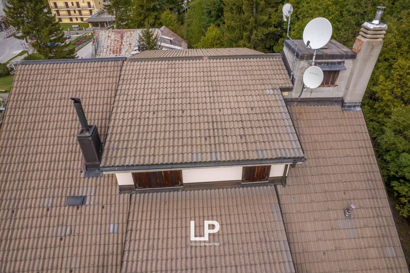 Top view of a tiled roof house, satellite dishes, chimney, and an elevated area, with a background of trees and other buildings