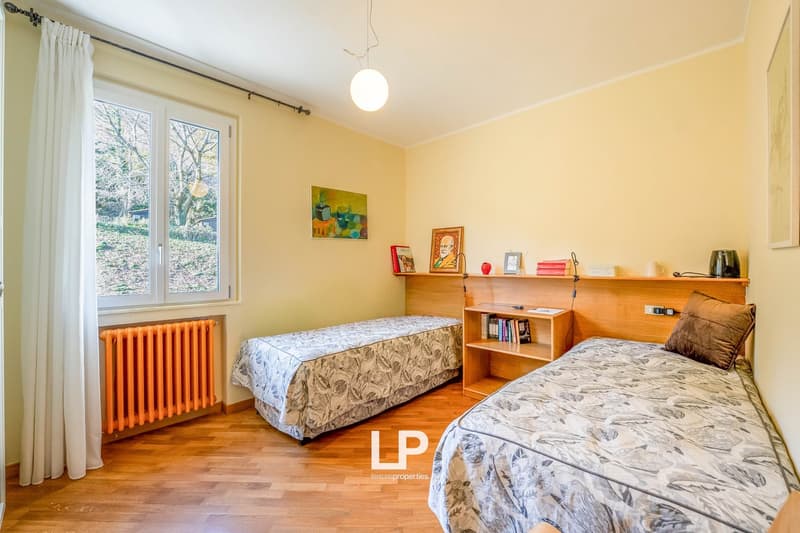 Double bedroom with two single beds, wooden headboards, wooden shelves, radiators, windows, curtains, floor lamp