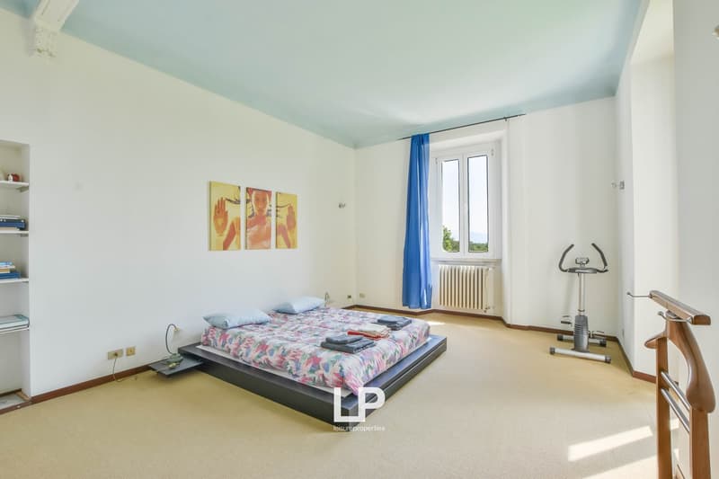 Bedroom with a bed, pillows, comforter, and nightstand with lamp. There is a window with a view, a shelf with books, and a white wall.