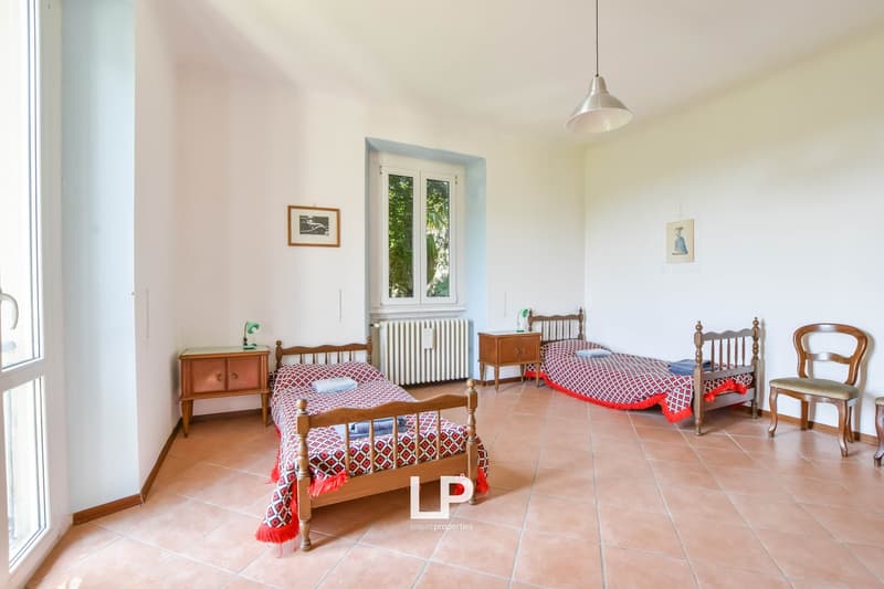 Bedroom with two single beds, a nightstand, a wooden cabinet, a white radiator, framed pictures on the wall, a lamp, and a window.