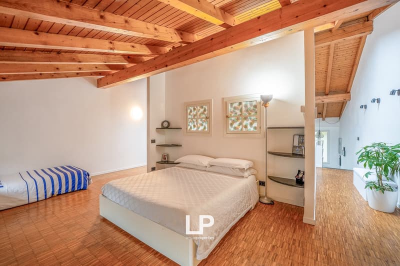 Minimalist bedroom with large bed, wooden floor, white walls, exposed wooden beams, nightstands, wall-mounted shelves, lamp, bed with white sheets and pillows, and an additional bed.