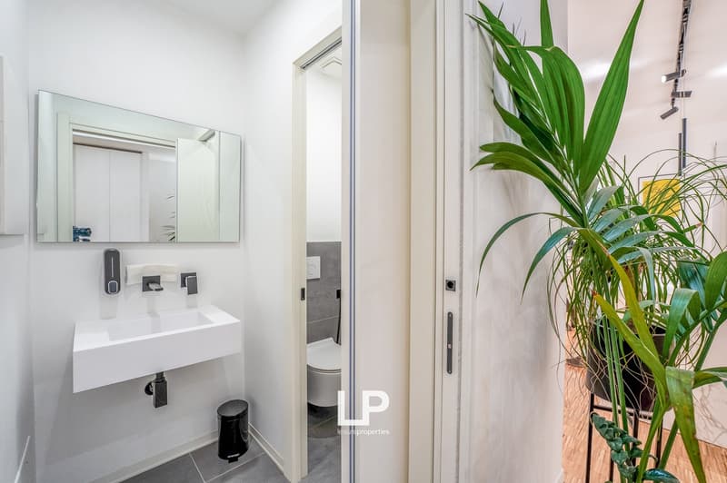 Modern bathroom with white sink, mirror, and grey tiled floor, toilet visible through the open door