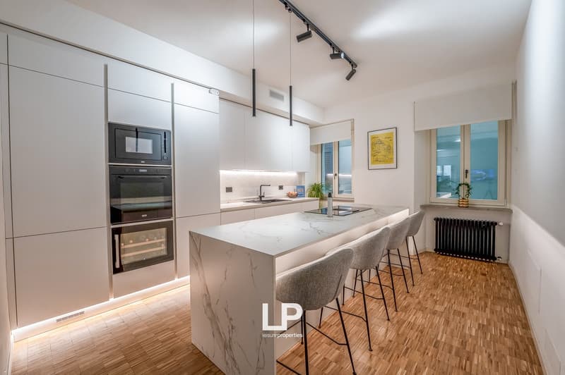 White cabinets, marble countertop, built-in microwave and oven, sink with faucet, stools, radiator, wooden floor, track lighting, window with view