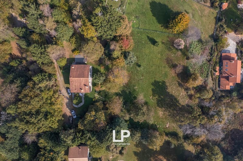 Aerial view of a house with a brown roof, nestled in a wooded area with multiple trees. The surrounding land is green with some trees having red leaves. A car is parked in front of the house.