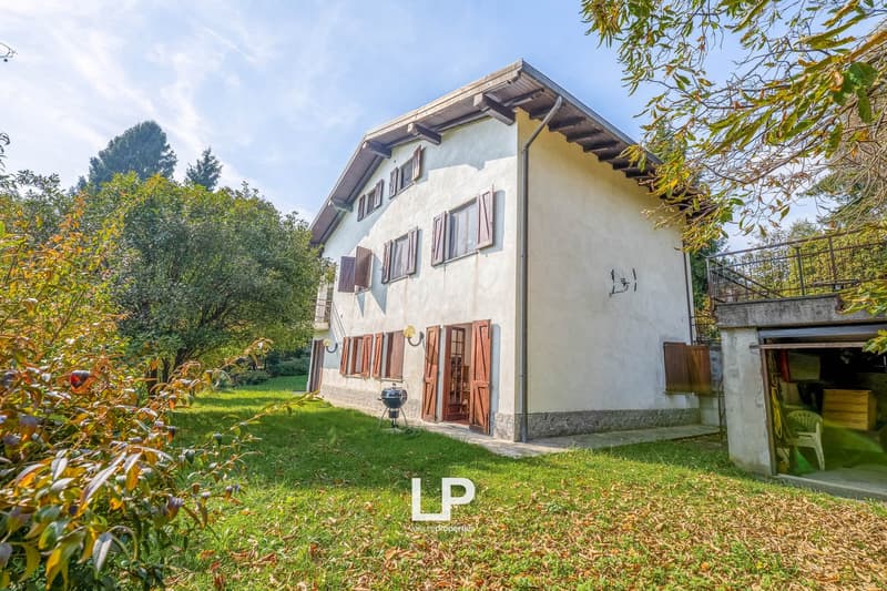 Two-story house with white exterior, wooden shutters, exposed wooden beams, brick foundation, open garage, balcony, green lawn, outdoor barbecue