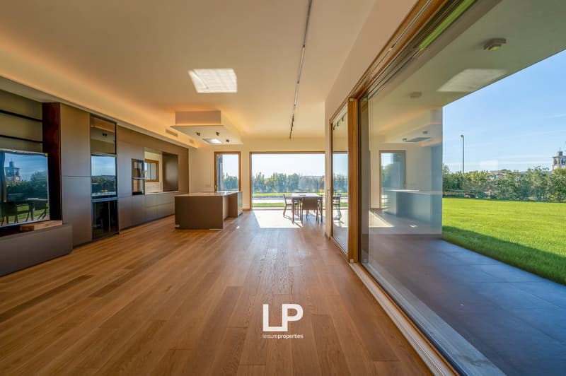 Large open space, glass doors, wooden floors, view of green lawn, table and chairs outside, kitchen area