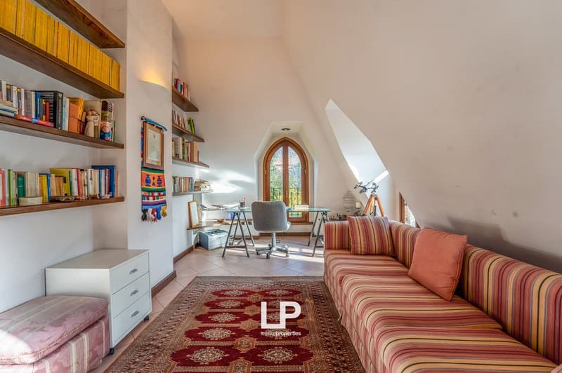 A spacious living room with an arched window, a desk with a chair, a couch, a rug, and shelves filled with books and decorative items
