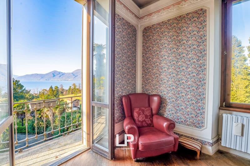 A room with a balcony featuring a metal railing, a red leather chair with a cushion, floral wallpaper, and a radiator. A window with a view of the sea, mountains, and greenery.