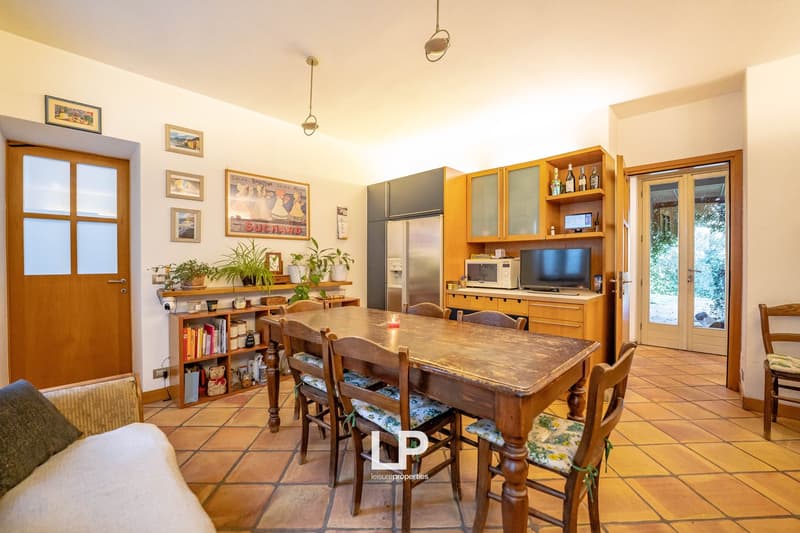 A dining area features a long dining table surrounded by wooden chairs. There is a large window leading to a patio and a shelf with books and plants. A couch is on the side and a microwave, television, and refrigerator are against the wall. A framed pictu