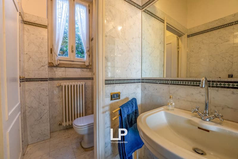 Modern bathroom with white marble tiles, white toilet, white sink, blue towel, radiator, window with white curtain, mirror