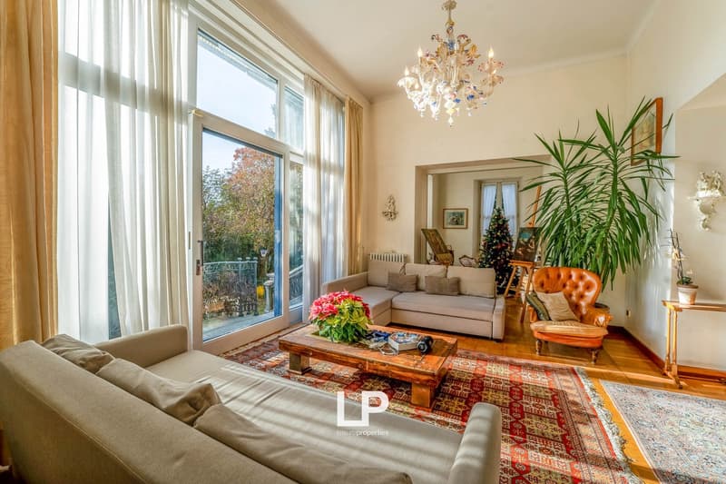 large windows, sliding doors leading to balcony, chandelier, oriental rug, couch, armchair, coffee table with flowers, picture frame