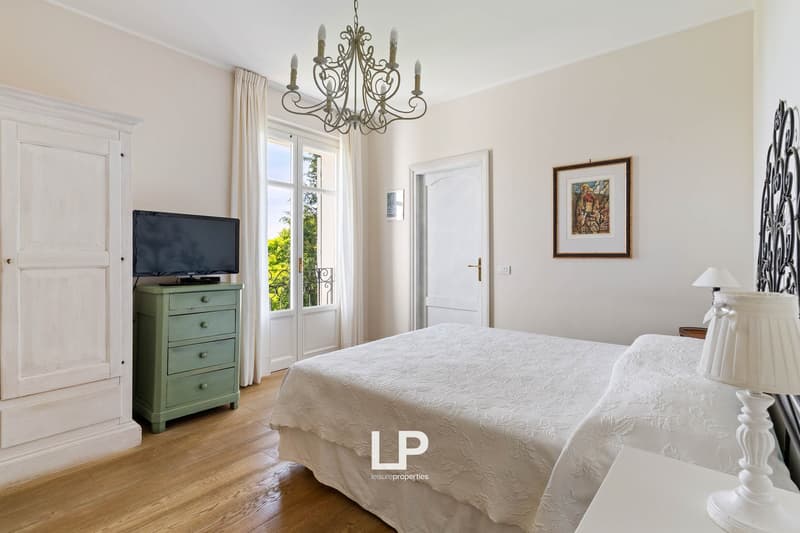 Spacious bedroom with wooden floor, large bed, dresser, bedside table, chandelier, white curtains, framed painting on the wall, lamp, TV on the dresser, balcony accessible through the window