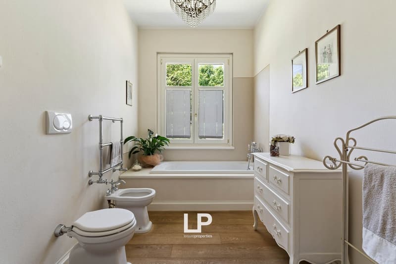 white bathtub, bidet, toilet, vanity, chandelier, white wooden floor, white walls, framed pictures, white cabinet with drawers