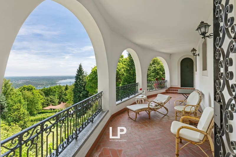 Terrace with a view, wrought iron railing, tiled floor, lounge chairs, table with flower vase, greenery in the background.